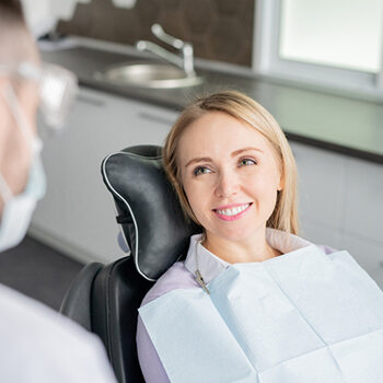 Happy Young Blonde Female Patient Looking At Her Dentist With Healthy Smile